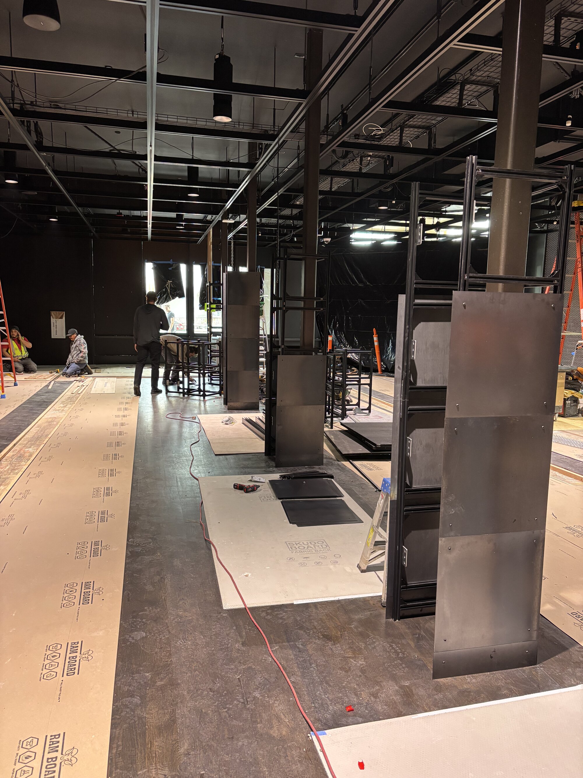 On-site installation — freestanding steel screen plinths going in across a raw buildout, floors masked with protection board